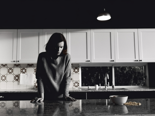 Frau steht mit gesenktem Blick in einer dunklen Küche und stützt sich auf eine Arbeitsfläche